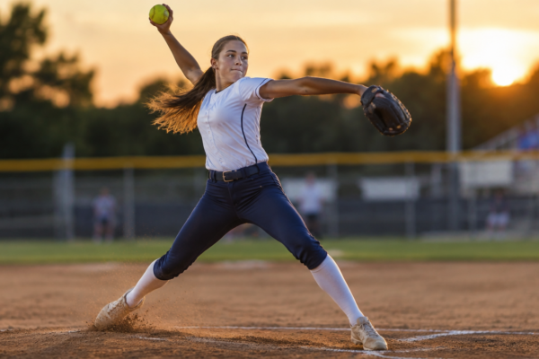 Softball pitching speed by age: teenage fastpitch pitcher driving off the rubber in her windmill delivery